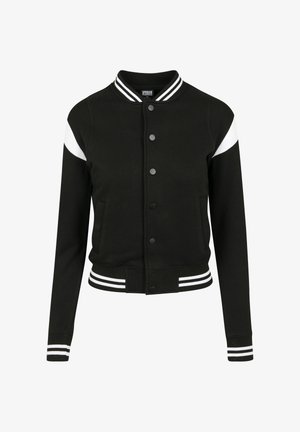 Czarna kurtka varsity z białymi paskami na ramionach, zapinana na guziki, z prążkowanymi mankietami i pasem; zestawiona z legginsami w kamuflażowy wzór.