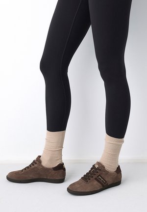 Legs wearing black leggings, beige ribbed socks, and brown suede sneakers with black soles standing on a white floor and wall background.