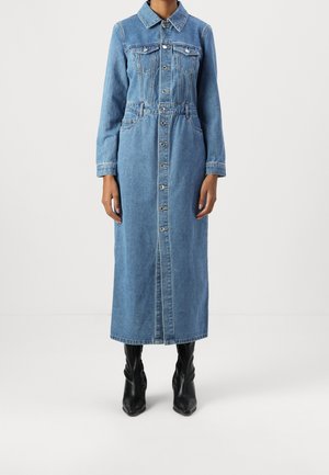 Vrouw draagt een lange blauwe denimjurk met knopen, lange mouwen, borstzakken en zwarte enkellaarsjes met spitse neus.