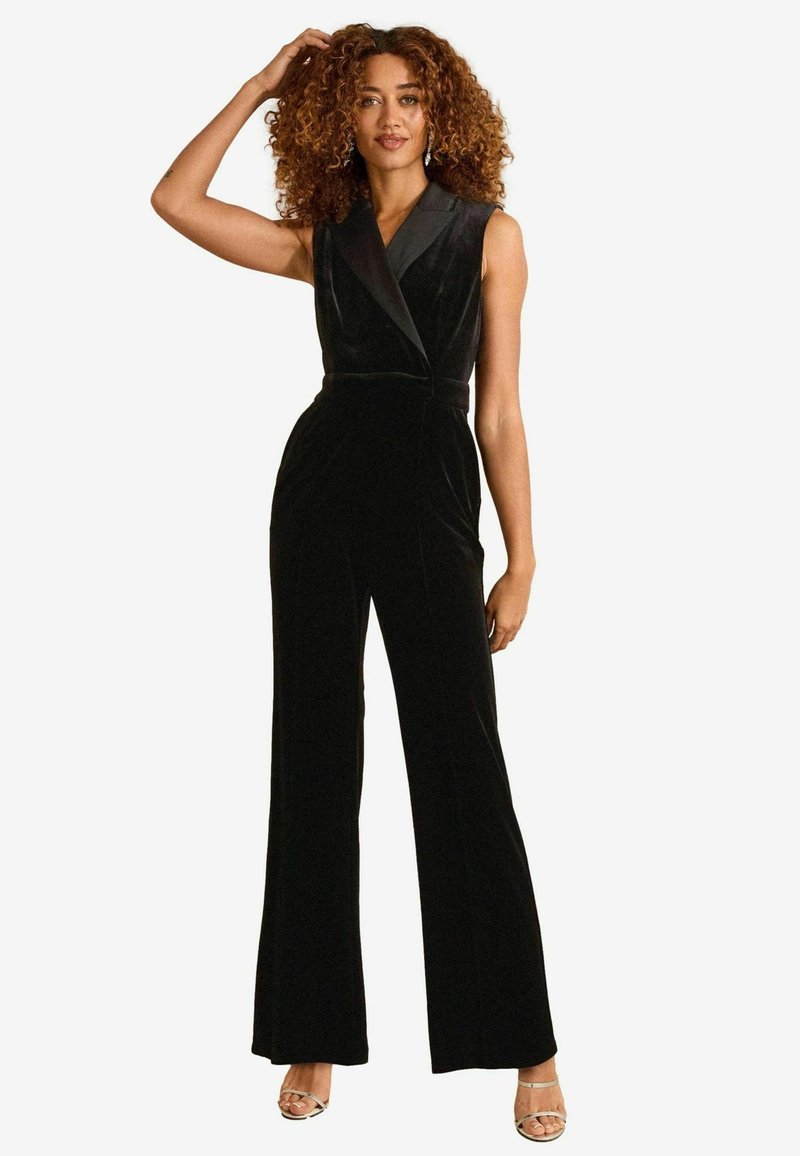 Salopetă din catifea neagră cu design fără mâneci, siluetă înfășurată, pantaloni largi și detalii contrastante la gulerul negru.