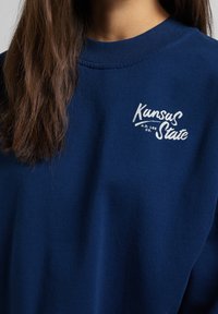 Sudadera azul marino con cuello redondo y detalles acanalados, con texto blanco que dice "Kansas State" y "H.D. Lee Co."