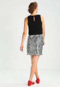 Mujer con cabello rizado, blusa sin mangas negra, falda geométrica en blanco y negro y zapatos de tacón bloque rojos caminando de espaldas sobre un fondo plano.