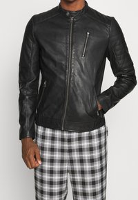 Svart läderjacka med hög krage, quiltade axeldetaljer, fickor med dragkedja framtill och en figurnära design, bärs över rutiga byxor.