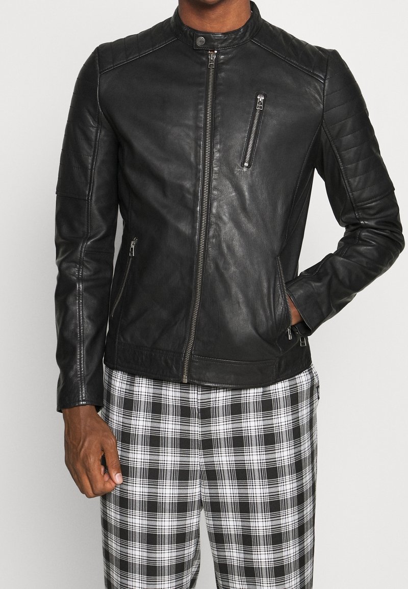 Svart läderjacka med hög krage, quiltade axeldetaljer, fickor med dragkedja framtill och en figurnära design, bärs över rutiga byxor.