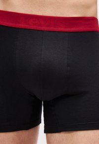 Μαύρα μπόξερ εσώρουχα με πλατιά κόκκινη μέση που φέρει διακριτική σήμανση της Levi's, φορεμένα από ένα άτομο από τη μέση έως τα μέσα των μηρών.