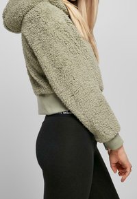 Grön fuskpälskort hoodie med huva, texturerad yta, elastisk nederkant, i kombination med svarta leggings med logoband i midjan.
