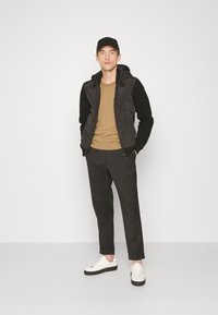 Jachetă cu glugă neagră, tricou maro, pantaloni închiși la culoare și adidași albi. Jacheta are mâneci texturate și două fermoare pentru închidere.
