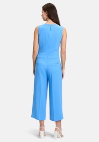 Completo blu senza maniche con scollo sulla schiena, gamba larga, due tasche sul retro e zip sul retro. Il tessuto ha una superficie liscia e opaca.