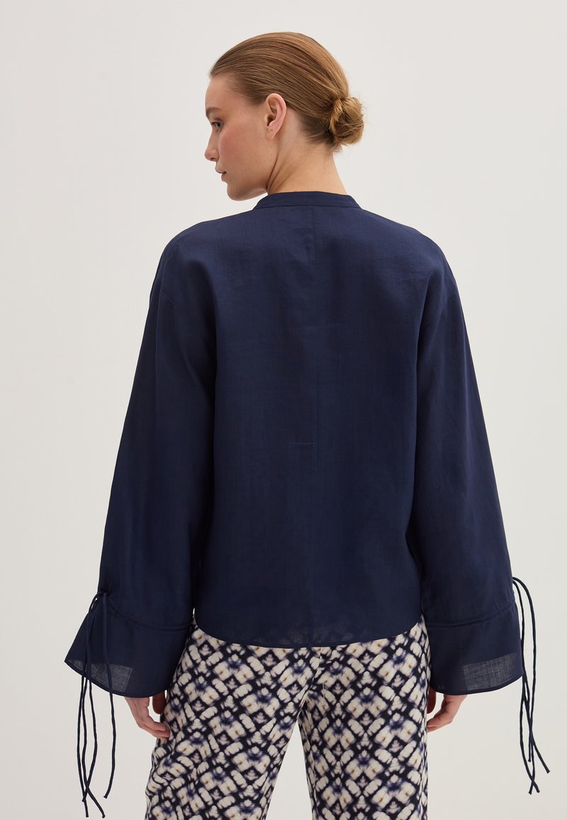Donna con i capelli raccolti in un bun, che indossa un ampio top a maniche lunghe blu navy con lacci ai polsini e pantaloni a gamba larga neri e bianchi a fantasia, vista da dietro.