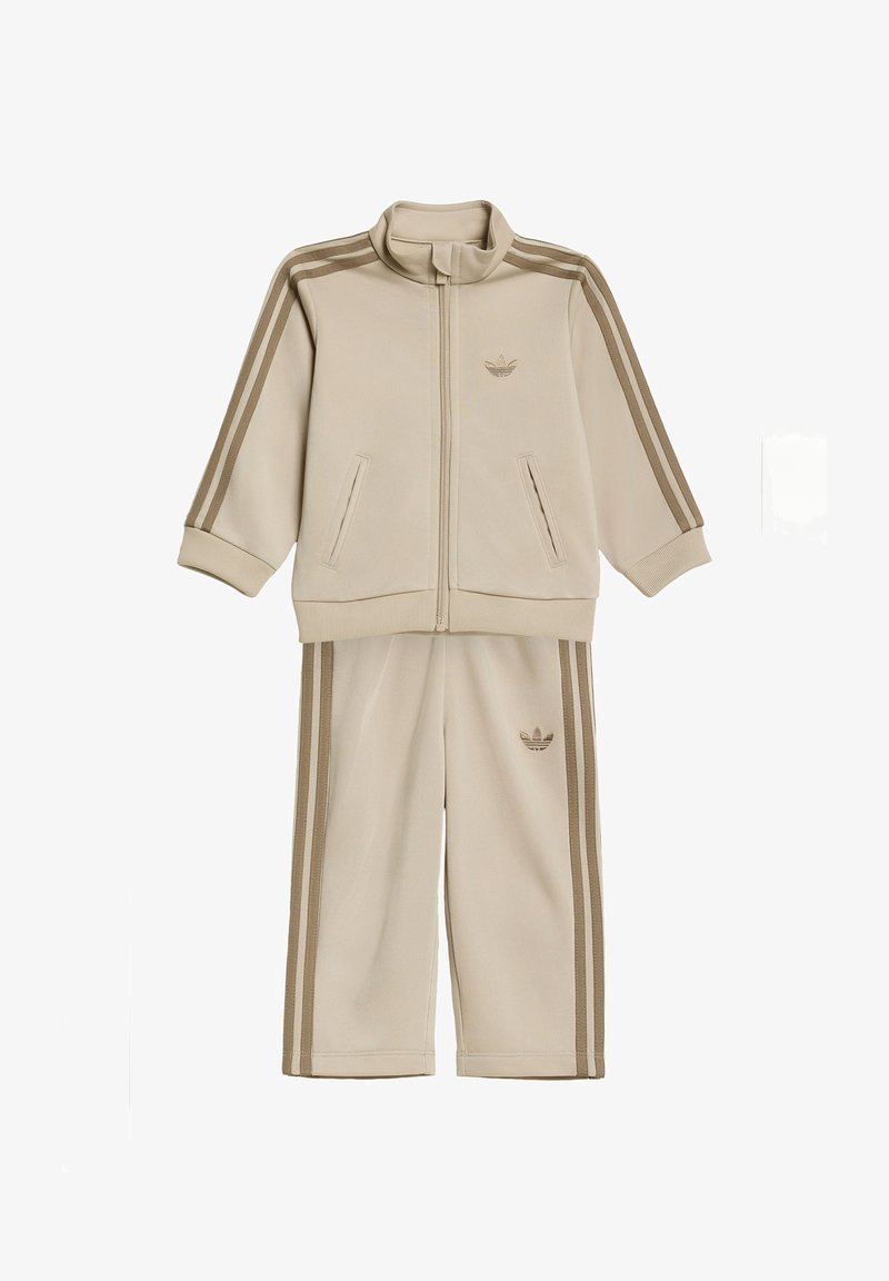 Survêtement beige zippé avec poignets côtelés, deux poches avant, et pantalon assorti avec double bande latérale et logo sur la poitrine et la cuisse.