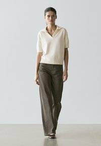 Elfenbeinfarbener Polo-Pullover mit kurzen Ärmeln und Kragen, kombiniert mit weit geschnittenen dunkelbraunen Hosen. Das Modell steht in einer minimalistischen Innenumgebung.