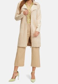 Beige mokkanahkainen takki lovikauluksella, yhdistetty kuvioidun keltaisen blousen, beigenväristen lyhyiden housujen ja vaaleanvihreiden korkokenkien kanssa.