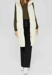 Gilet lungo imbottito di colore crema con cappuccio, chiusura con zip e cuciture a contrasto, indossato sopra una maglietta verde e pantaloni neri a vita alta.