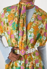 Farverig floral-mønstrede omslag med grønne, orange, pink og blå nuancer, parret med en lyseblå plisseret top og en hvid bælte i taljen.