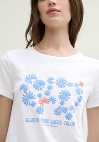 Hvit bomull t-skjorte med blå og røde blomsterdesign og teksten "BLÅ ER DIN LYKKEFARGE" i blått, rund halslinje, korte ermer.