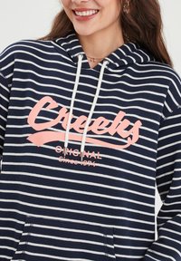 Felpa a righe blu navy e bianco con cappuccio e cordino, dotata di un logo rosa "Creeks" e la scritta "Original Since 1974". Include una tasca frontale.