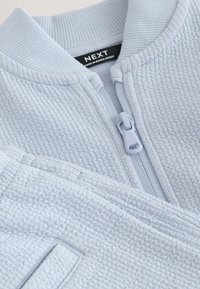 Light gray textured zip-up garment with ribbed collar and visible black label reading "NEXT made in Bangladesh."