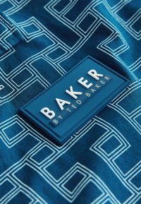 Niebieska tkanina z białym geometrycznym wzorem kwadratów, z niebieską prostokątną naszywką opisaną "BAKER BY TED BAKER" wypukłymi białymi literami.