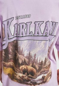 Purple cotton t-shirt featuring "Karl Kani" text and a graphic of a bear in a forest landscape, with detailed trees and rocky terrain.
