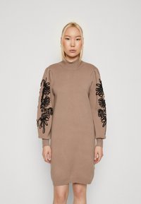 Strikket kjole i beige med høj hals, med sort blomstermotiver på puffede ærmer og ribbede manchetter. Blød tekstur, knælangt design.