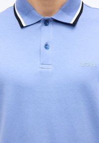 Polo bleu clair avec deux boutons bleus, col texturé à rayures noires et blanches, et "BOSS" brodé sur la poitrine.