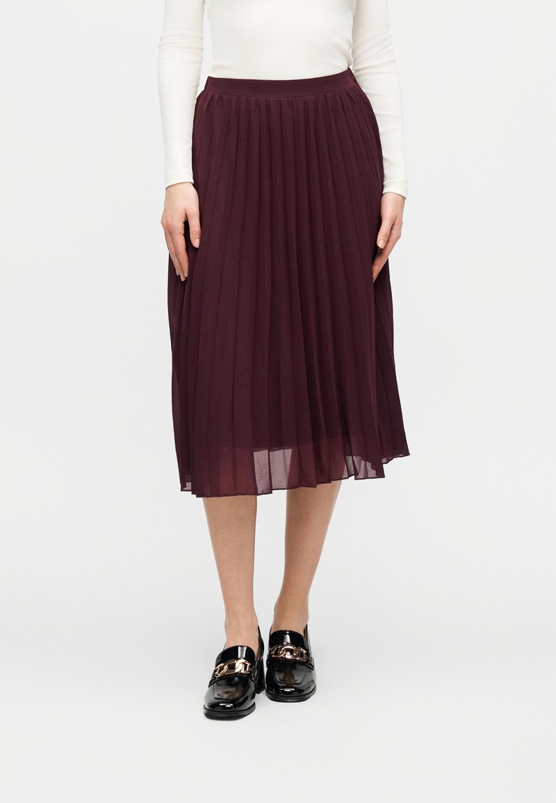 Femme portant un haut blanc à manches longues rentré dans une jupe midi plissée bordeaux taille haute, accompagnée de mocassins noirs ornés d'une chaîne dorée.