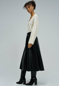 Femme aux cheveux bouclés portant des boucles d'oreilles en or, un pull blanc à col en V, une jupe midi noire texturée et des bottes à talons noires, se tenant de côté.