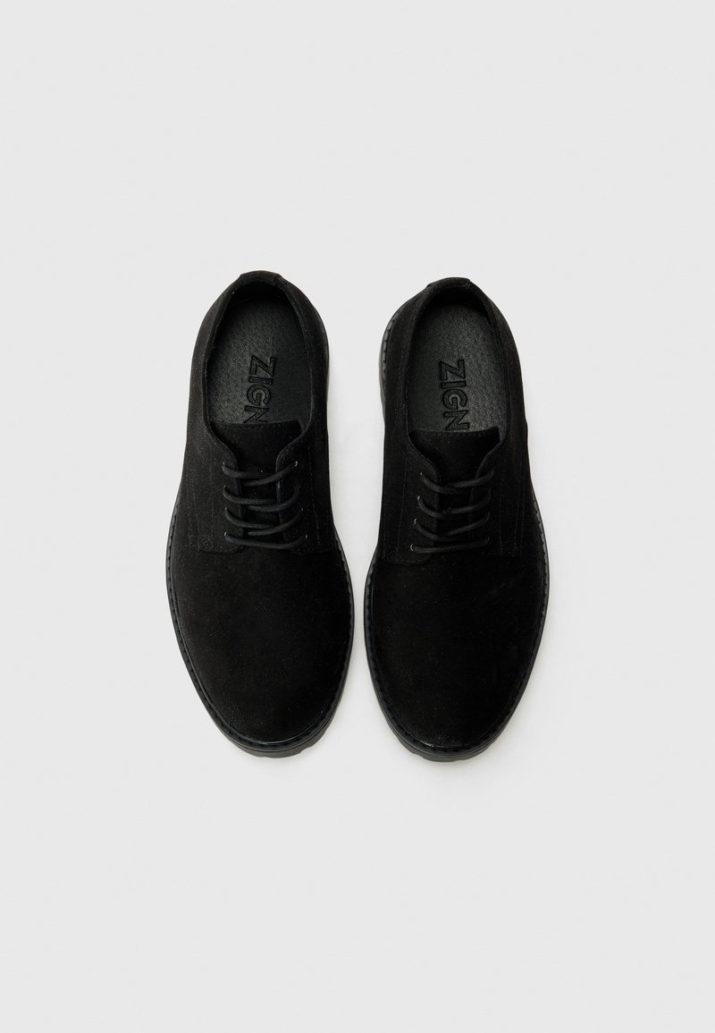 Chaussures en suede noir avec une semelle plate, un bout rond et un design à lacets. Un style minimaliste qui présente une accent texturé au niveau de la languette et une marque sur la semelle intérieure.