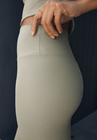 Leggingsid pehmest, heledast rohelisest kangast, millel on sile tekstuur, kõrge vöökoht ja külgmised õmblused, osaliselt nähtav käsi vöökohal.