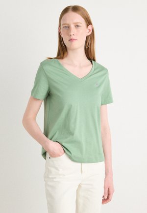 Green v-neck t-shirt made of soft fabric with short sleeves. Features a subtle logo on the chest, paired with light-colored pants.