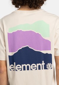 Cremfarbiges T-Shirt mit einem geschichteten Berggrafik in Grün, Lila und Marineblau, mit dem Wort "Element" und Logo in fettem Weiß gedruckt.