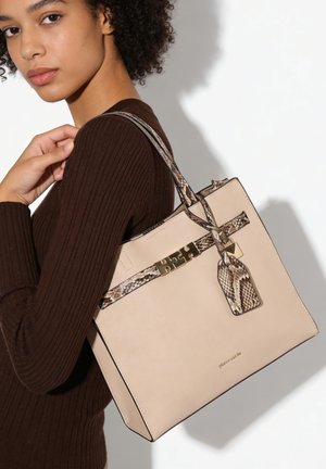 Jonge vrouw in een bruine geribde trui draagt een beige shopper met slangenprintaccenten en gouden hardware over haar schouder.