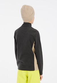 Fleece-Pullover mit hohem Kragen, schwarzer Rückseite und beigen Seiteneinsätzen. Kombiniert mit einer gerippten, beigen Mütze und leuchtend gelben Hosen.