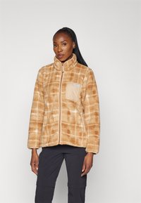 Veste en polaire beige avec une texture douce, présentant un motif en grille, un devant zippé et une poche latérale. Comprend un col montant pour plus de chaleur.