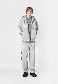 Sudadera gris con cremallera y logo blanco, superpuesta a una camiseta gris. Pantalones de chándal grises a juego con bolsillos. Zapatos negros texturizados.