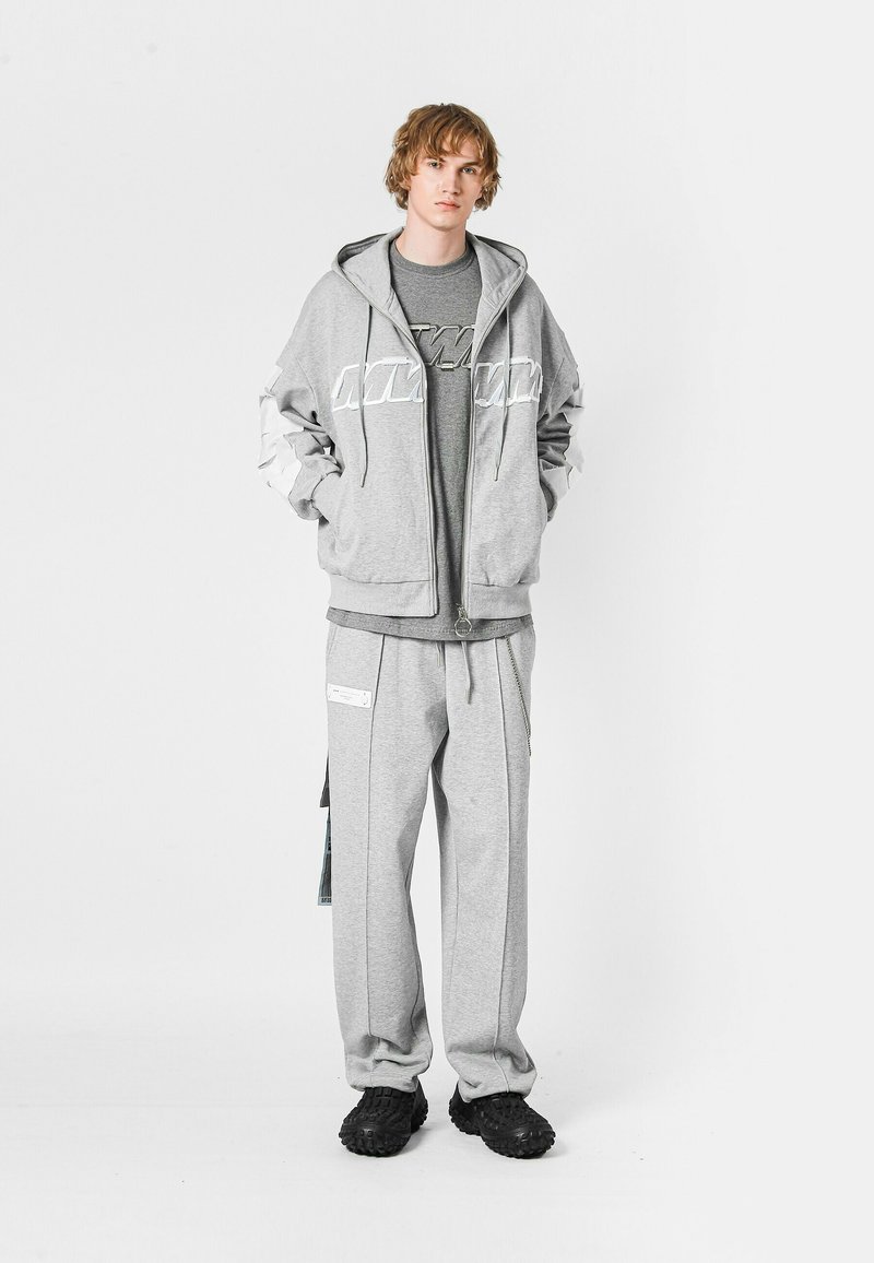 Sudadera gris con cremallera y logo blanco, superpuesta a una camiseta gris. Pantalones de chándal grises a juego con bolsillos. Zapatos negros texturizados.