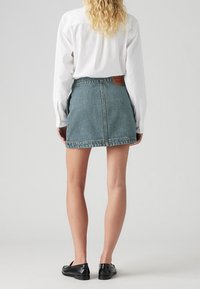 Falda de mezclilla en verde azulado con corte recto, con una cintura de altura media y etiqueta en la parte posterior. Combinada con una camisa blanca de manga larga y zapatos negros.