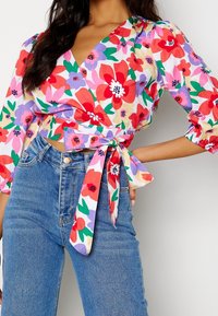 Un blouse à manches bouffantes avec un motif floral présente des fleurs rouges, roses et violettes sur un fond blanc, avec un lien à la taille. Porté avec un jean bleu.