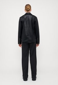 Svart skinnjakke med skulderklaffer, lange ermer og glidelås på mansjettene, kombinert med svarte pinstripede bukser og kraftige svarte støvler.