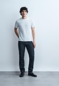 Mladý muž s kučeravými vlasmi stojaci pri bielej stene, oblečený v bielom tričku s pruhmi, tmavých džínsoch a čiernych mokasínach, ruky uvoľnené.