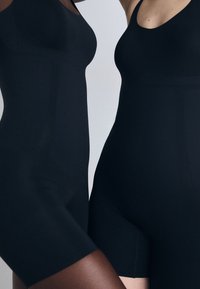 Black bodysuits made of smooth fabric, featuring a scoop neck and short legs. The design is minimalist with no visible hardware or patterns.