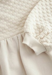 Kermavärinen mekko, jossa on tekstuuri kudottu yläosa, laskostettu hame ja ribbauskut. Pehmeä kangas, jolla on sileä, kevyen näköinen ilme.