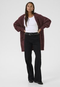 Maroon knitted cardigan over a white shirt, paired with black flared jeans and a black belt. Smooth textures and relaxed fit.