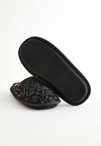 Chaussons tricotés noirs et gris avec une dessus texturé, une forme ronde, et une semelle en caoutchouc, avec une petite étiquette sur le côté.