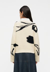 Γυναίκα με μακριά σκούρα μαλλιά που φοράει εκρού πουλόβερ με μαύρα αφηρημένα λουλουδάτα σχέδια και σκούρα φούστα από τζιν, γυρισμένη προς τα πίσω.