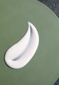 White cream swatch on a textured green plate. The cream has a smooth, glossy finish and a curved, elongated shape.