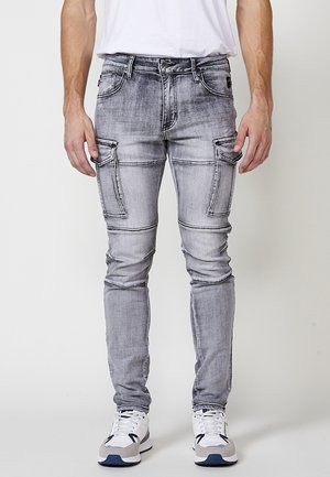 Gray distressed cargo jeans with multiple side pockets, fitted design, and contrast stitching. Paired with white sneakers.