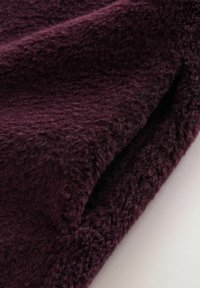 Hladká, hlboká burgundská tkanina s chlpatej textúrou, jemne zhromaždená na okraji, ktorá predvádza hustý, mäkký vzhľad.