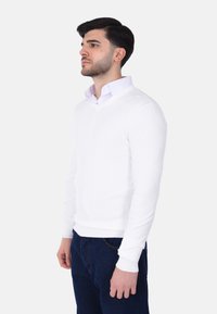 Giovane uomo che indossa un maglione bianco sopra una camicia bianca con colletto e jeans blu scuro, in piedi e che guarda verso sinistra.