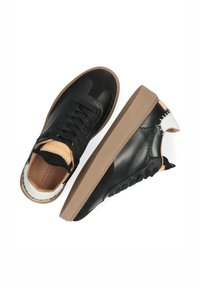 Par de zapatillas negras de caña baja con suelas marrones, acentos blancos en el talón, cordones negros y forro interior marrón claro, mostradas sobre un fondo blanco.